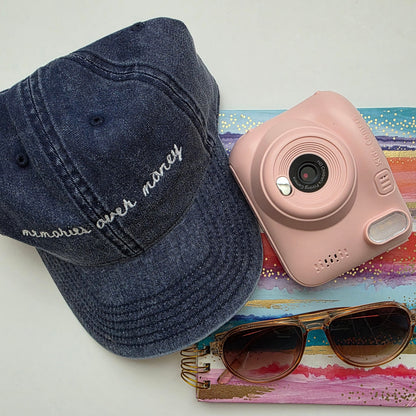 Navy cap with 'memories over money' text, pink camera, sunglasses, and colorful book on a white surface