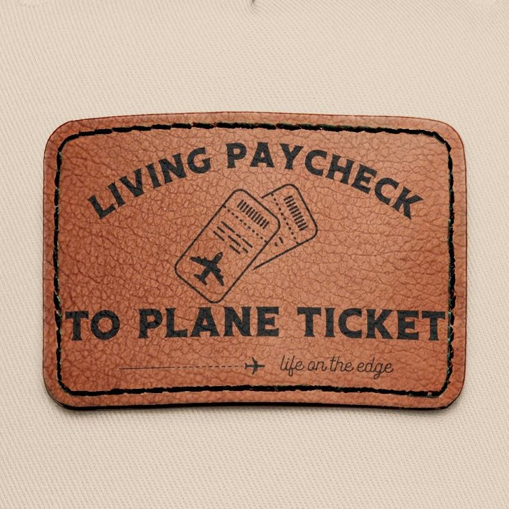 Beige and green cap with a brown leather patch displaying 'Living Paycheck to Plane Ticket"