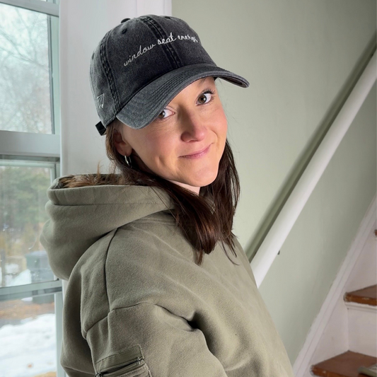 Person wearing a green hoodie and black travel cap with "window seat energy indoors