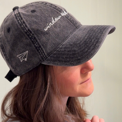 Person wearing a vintage wash black cap with 'window seat energy' embroidery on a plain background