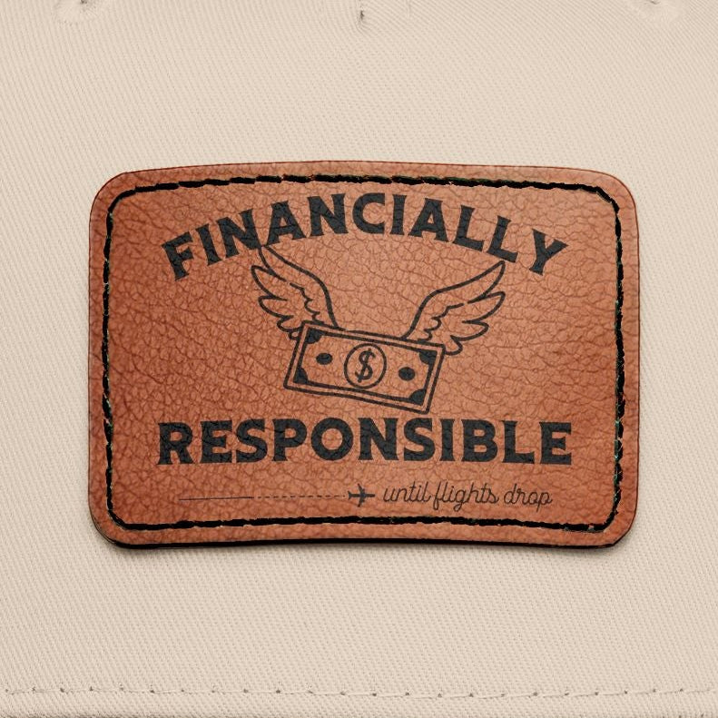 A two-tone baseball cap with a cream-colored crown and a black curved brim, featuring a brown faux-leather patch on the front that reads 'FINANCALLY RESPONSIBLE (until flights drop)'