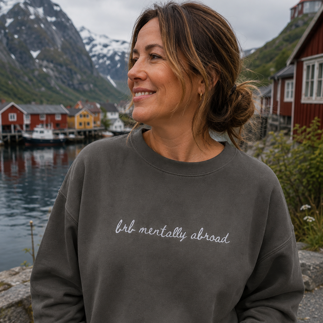 A woman wearing a dark gray crewneck sweatshirt with the phrase 'brb mentally abroad' written in white script across the chest, posing outdoors with a scenic backdrop of mountains, water, and red houses.