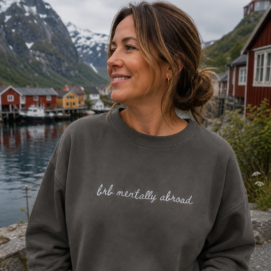 A woman wearing a dark gray crewneck sweatshirt with the phrase 'brb mentally abroad' written in white script across the chest, posing outdoors with a scenic backdrop of mountains, water, and red houses.