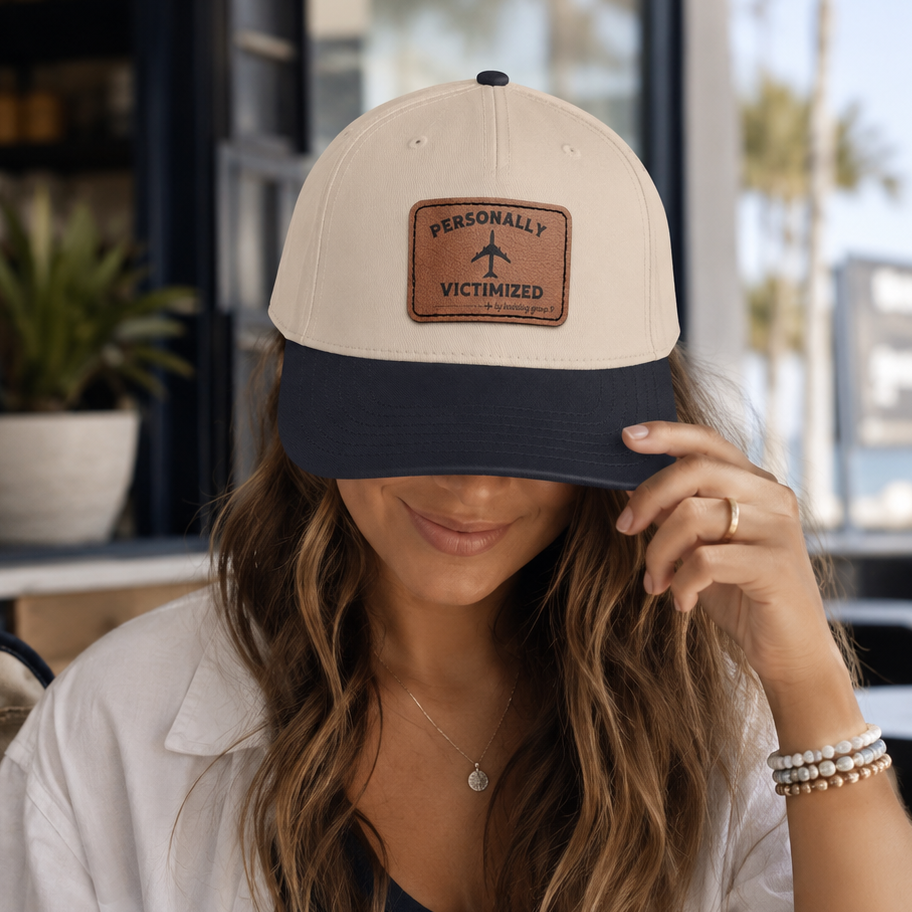 A woman in a white shirt and navy top wears a beige and navy baseball cap with a brown patch reading 'PERSONALLY VICTIMIZED' and an airplane icon, sitting at a cafe table with a coffee cup nearby.