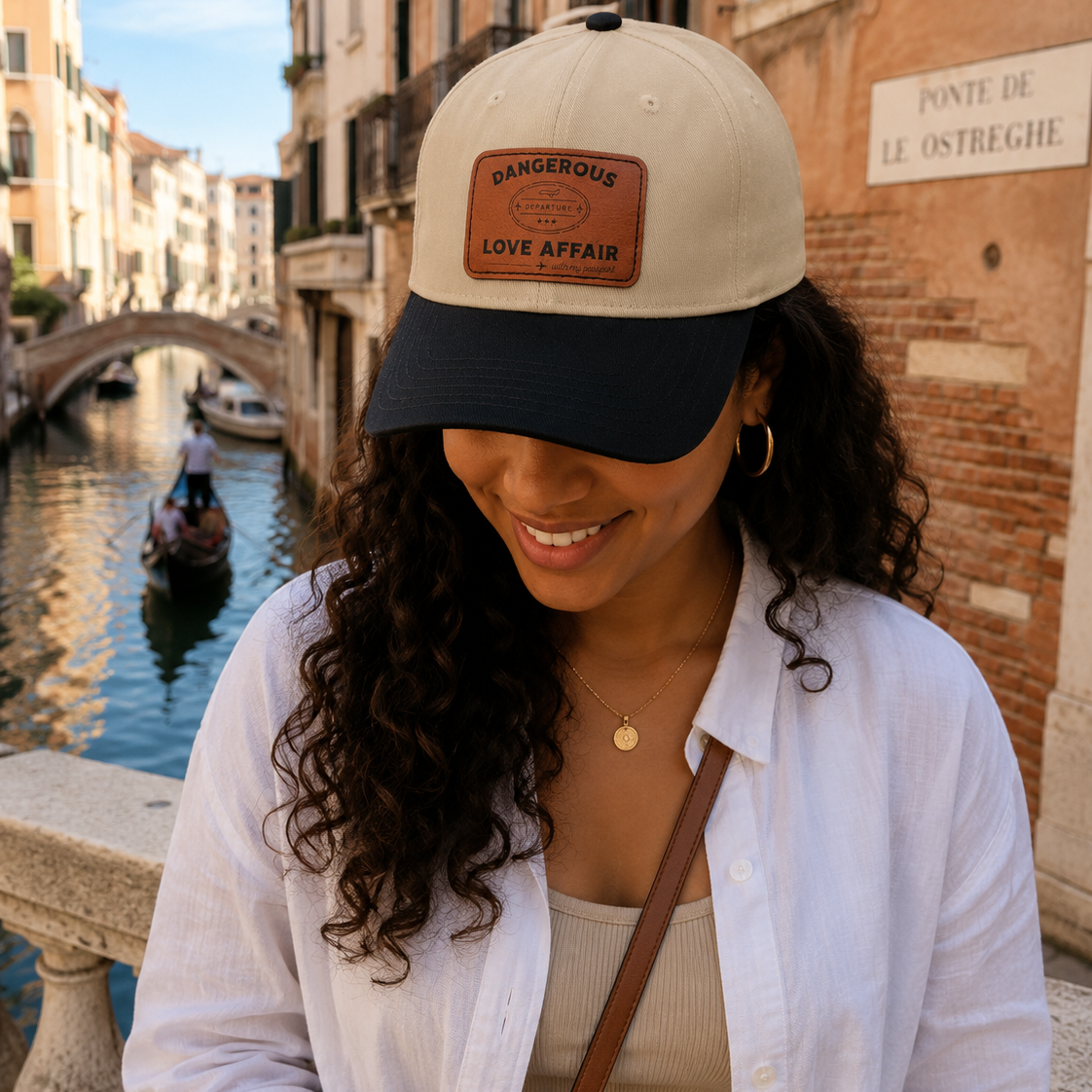 A woman wearing a beige and navy blue baseball cap with a brown leather patch that reads 'DANGEROUS LOVE AFFAIR (with my passport)' stands by a canal in Venice, with a gondola and historic buildings in the background.