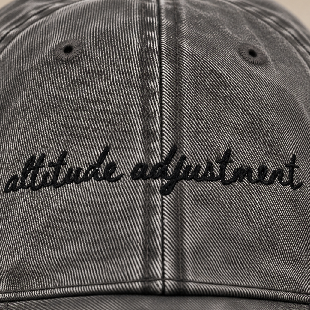 Close-up of a washed black baseball cap with 'altitude adjustment' embroidered in black script across the front.